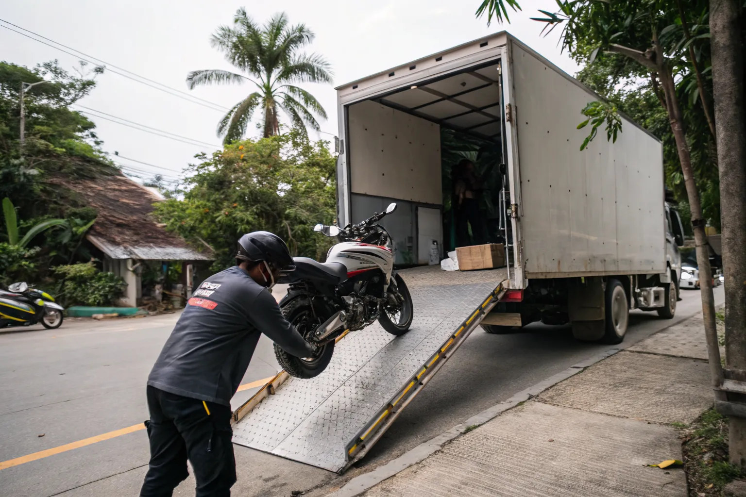 Crew member rolling a large adventure motorcycle up an aluminium ramp into a box truck on a Thai street